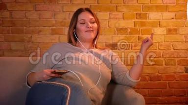 超重的长发女<strong>模特</strong>坐在沙发上，在舒适的家庭氛围中，用<strong>耳机</strong>听音乐。